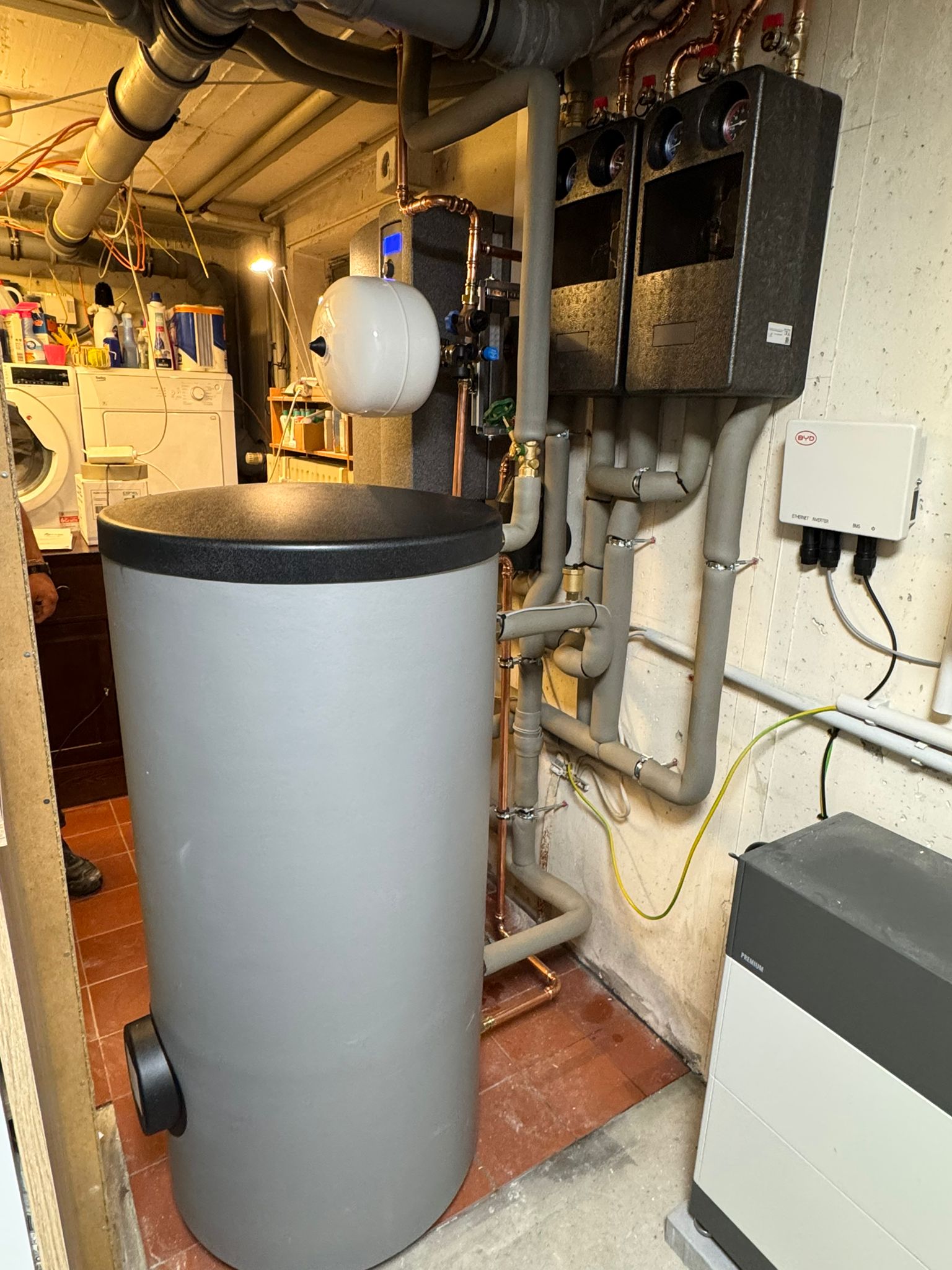 Technikraum mit Pufferspeicher, Wandgerät, zwei Ausdehnungsgefäßen und zahlreichen Rohrverbindungen.